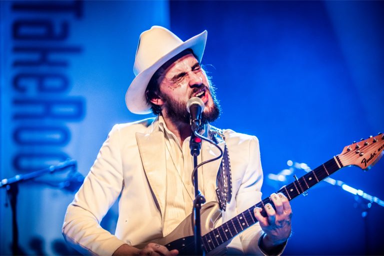 Een muzikant in een wit pak en hoed speelt gitaar en zingt op een podium met blauwe verlichting.