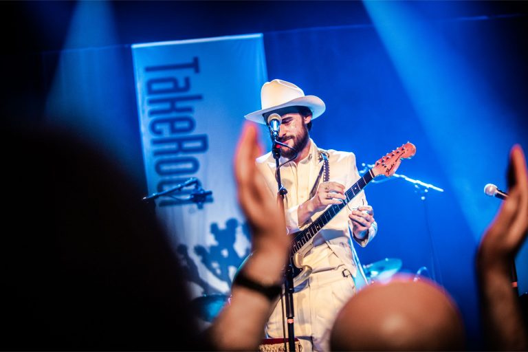 Een muzikant in een witte outfit en hoed speelt gitaar op een podium, met een banner op de achtergrond met de tekst 