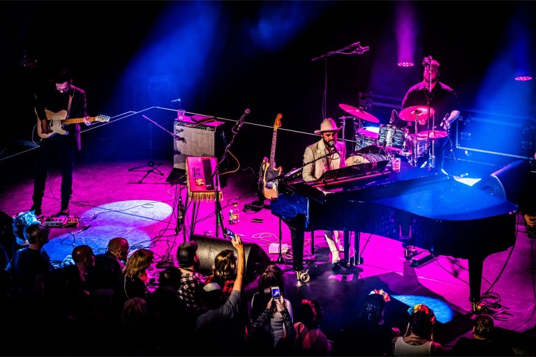 Een muzikant speelt piano op het podium terwijl een drummer en een publiek in de achtergrond zich genieten van het optreden onder gekleurde lichten.