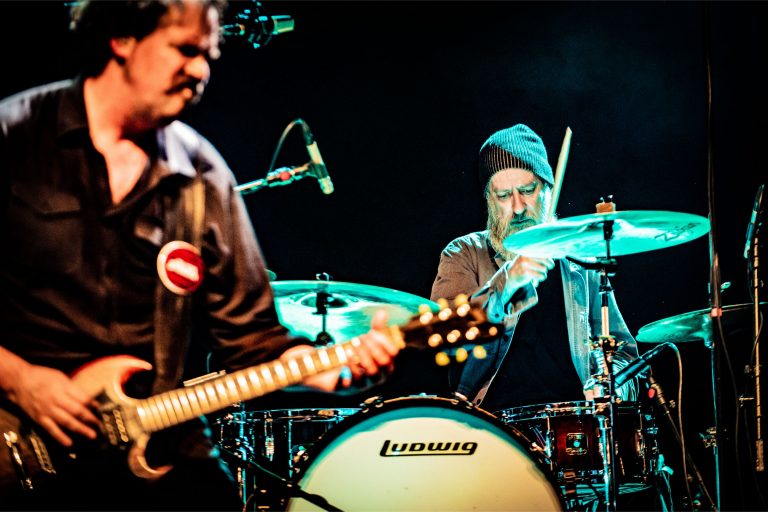 Twee muzikanten spelen samen op het podium, met de focus op een gitarist en een drummer.