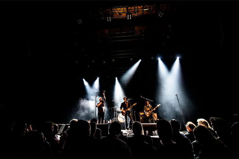 Een band speelt op een podium met sterke lichtstralen en een publiek dat kijkt.