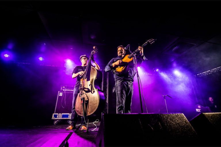 Twee muzikanten spelen op een podium met paarse verlichting, één met een gitaar en de ander met een contrabas.