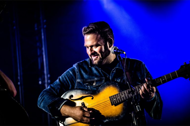 Een muzikant speelt met een gitaar op een podium met blauwe lichteffecten.