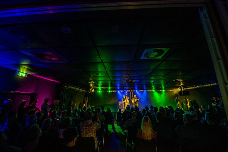 Een drukke concertzaal met kleurrijke verlichting en een optreden op het podium.