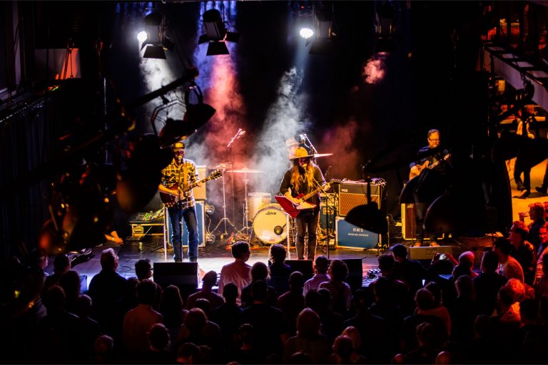 Een band performt op een podium met kleurrijke lichten en een enthousiast publiek ervoor.