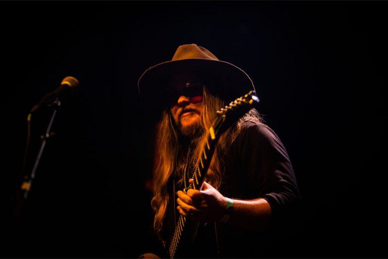 Een muzikant met lang haar en een grote hoed speelt gitaar op een donkere achtergrond.