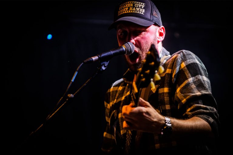 Een muzikant in een zwart-wit geblokte shirt speelt gitaar en zingt in een microfoon op een podium.
