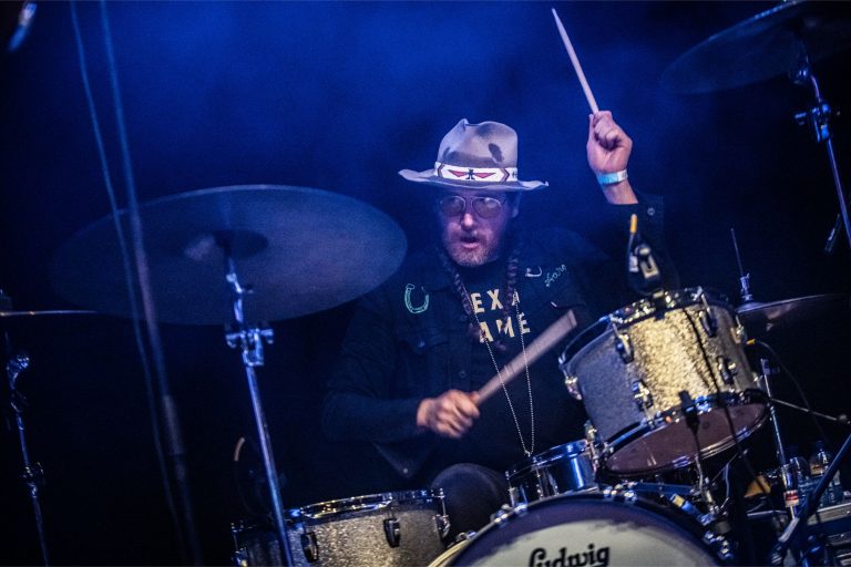 Een drummer speelt enthousiast op zijn drumstel terwijl hij een hoed draagt en omringd is door blauwe lichten.