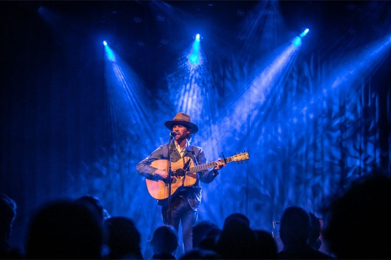 Een muzikant speelt gitaar op een podium, omringd door blauwe lichtstralen en een publiek.