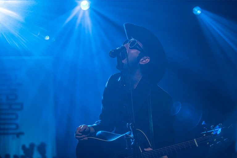 Een muzikant zingt en speelt gitaar op het podium, omringd door blauwe lichtstralen.