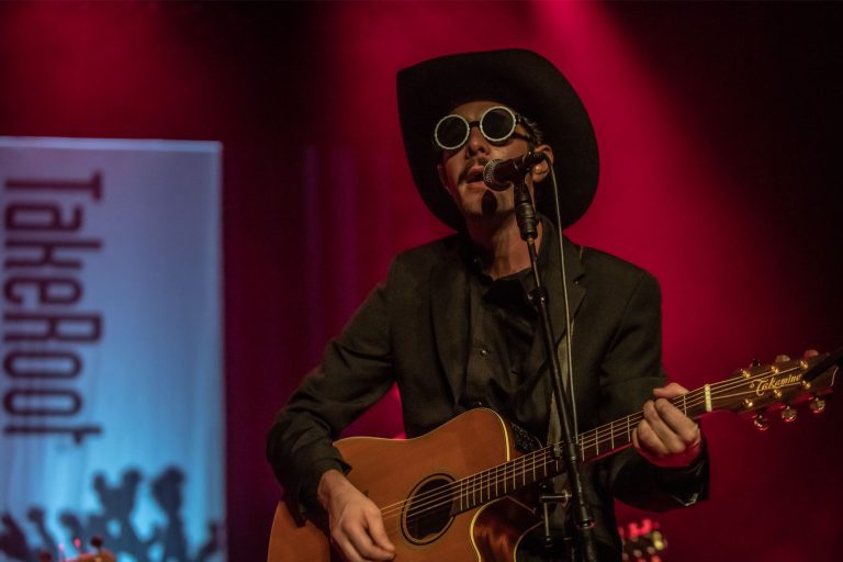 Een muzikant met een hoed en zonnebril speelt gitaar op een podium met rode belichting.