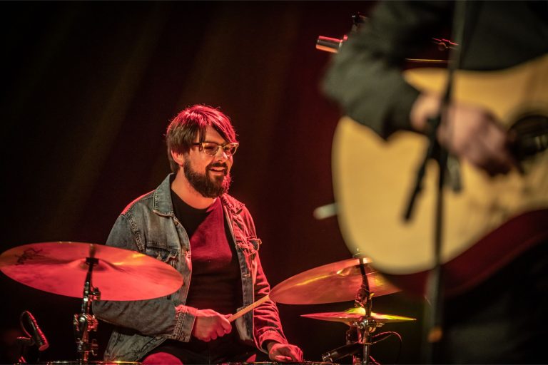 Een drummer in een spijkerjack zit achter zijn drumstel met een gitaarist op de voorgrond.