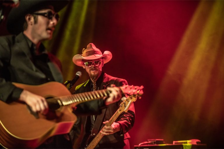 Twee muzikanten optreden op een podium met gekleurde lichten, de een met een gitaar en de ander met een hoed en zonnebril.