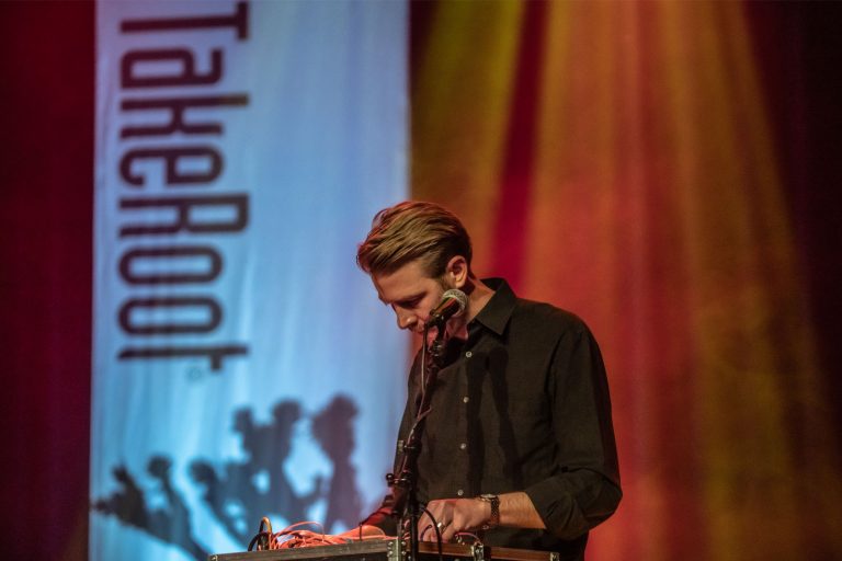 Een muzikant speelt op een keyboard voor een kleurrijke achtergrond met silhouetten.