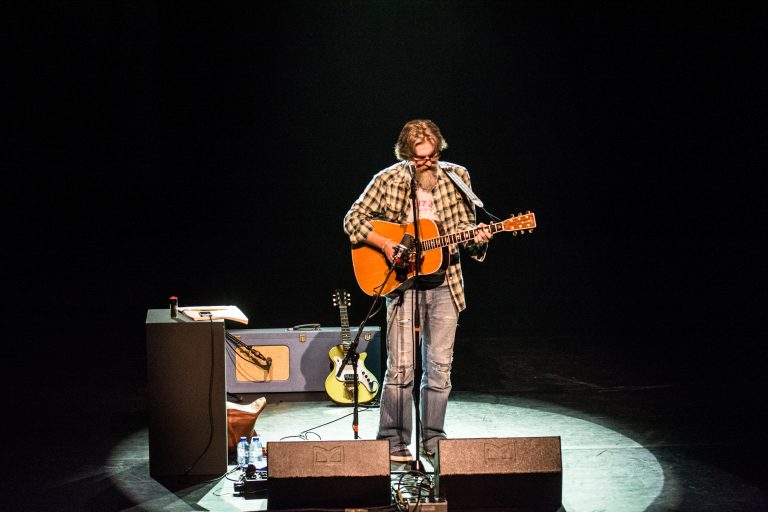 Een muzikant met een gitaar zingt op een podium, omgeven door een donkere omgeving.