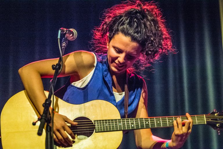 Een muzikante speelt een akoestische gitaar op het podium met een stralende glimlach, omringd door kleurrijke verlichting.