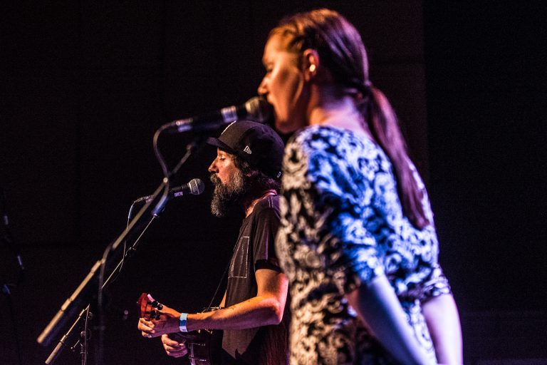 Een man speelt gitaar en zingt terwijl een vrouw op de achtergrond zingt in een donkere setting.