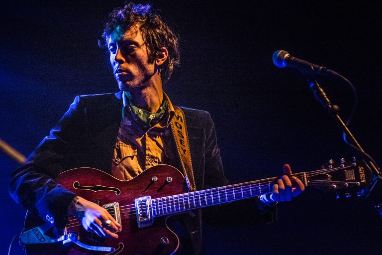 Een muzikant met krullend haar die een elektrische gitaar bespeelt op een podium met blauwe verlichting.