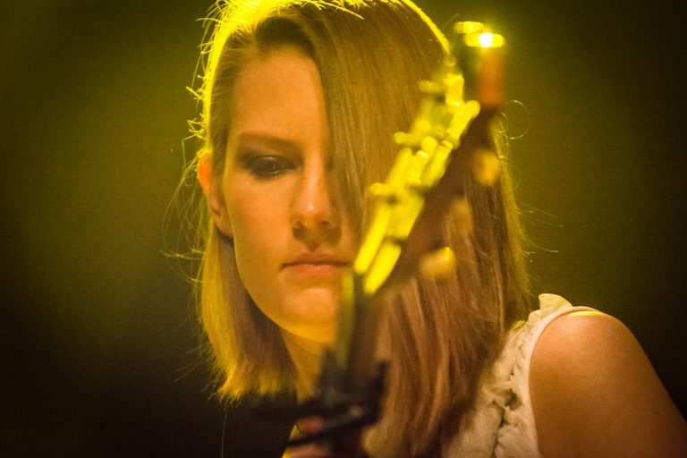 Een vrouw speelt gitaar met een intense blik, omringd door een warme, geelachtige verlichting.