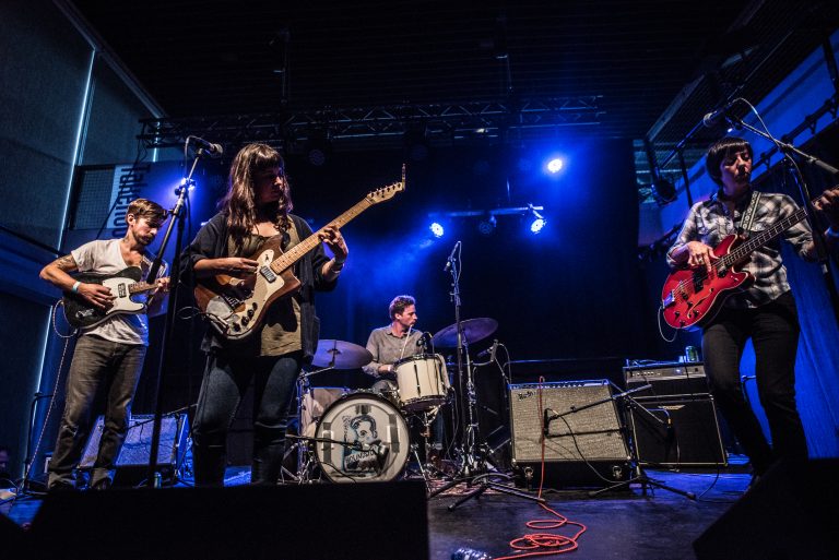 Een band speelt live op een podium met blauwe verlichting, waaronder een gitarist, een drummer en een andere muzikant met een elektrische gitaar.