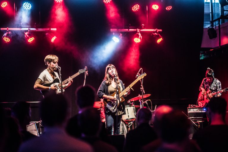 Een band staat op een podium met kleurrijke verlichting, terwijl ze muziek spelen voor een publiek.
