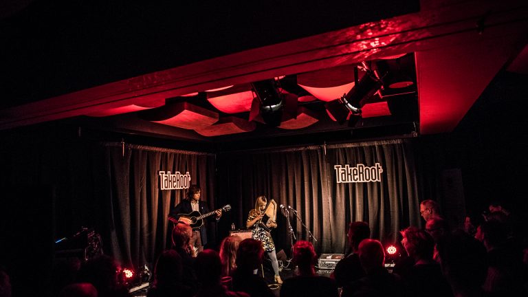 Een muzikale live-optreden in een sfeervol verlichte ruimte met een zangeres en een gitarist op het podium.