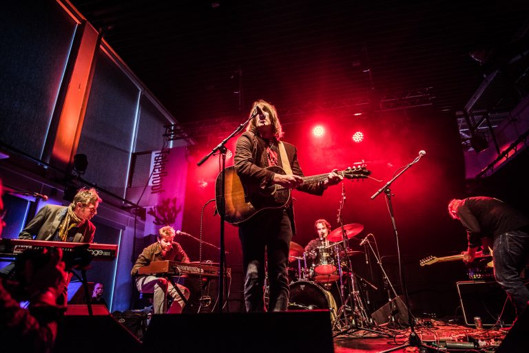 Een band speelt live op een podium met rode verlichting, waarbij de zangeres gitaar speelt en omringd is door muzikanten.