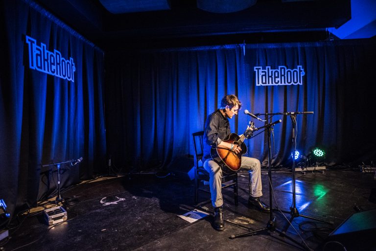 Een muzikant speelt gitaar op een podium met blauwe verlichting en een zwarte achtergrond.