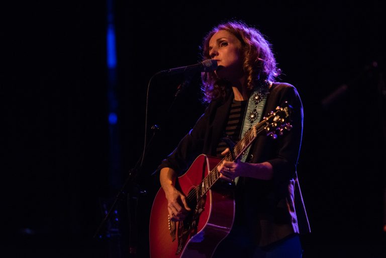 Een zangeres met krullend haar speelt gitaar en zingt op een podium met sfeervolle verlichting.