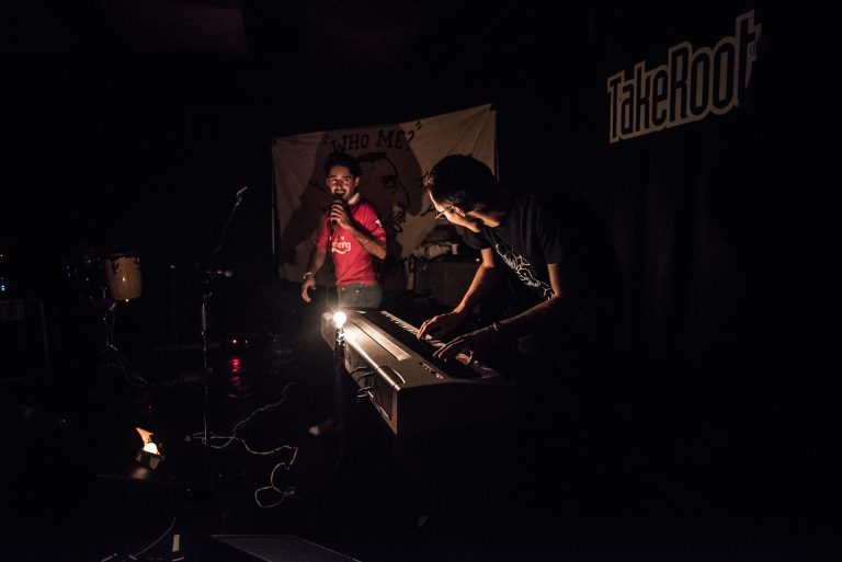 Twee muzikanten optreden op een donker podium, met één muzikant aan een keyboard en de ander met een microfoon.