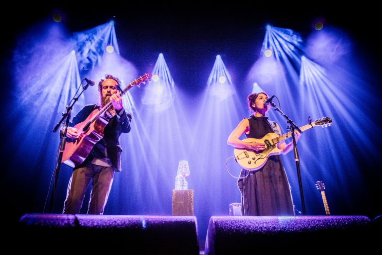 Twee muzikanten staan op een podium, met instrumenten in de hand en blauwe lichten die de achtergrond verlichten.