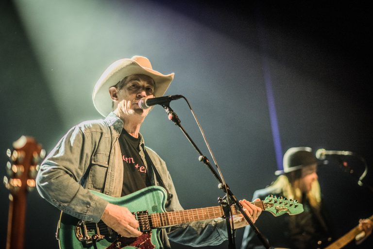 Een muzikant met een cowboyhoed speelt gitaar en zingt in een podiumomgeving met lichteffecten.