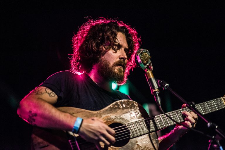 Een muzikant met krullend haar speelt op een gitaar, terwijl hij geconcentreerd naar zijn instrument kijkt.