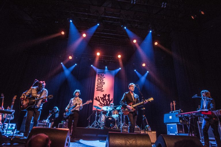 Een band voert live op met kleurrijke lichten op het podium en een spandoek op de achtergrond.