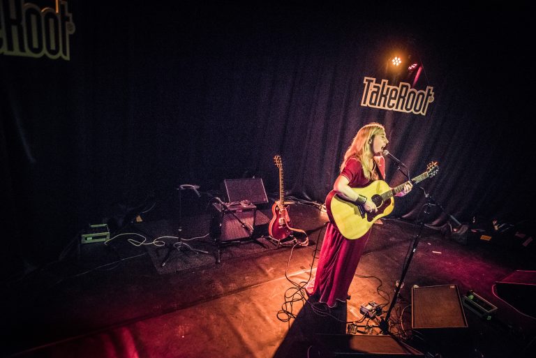 Een muzikante in een rode jurk staat op het podium met een gitaar.