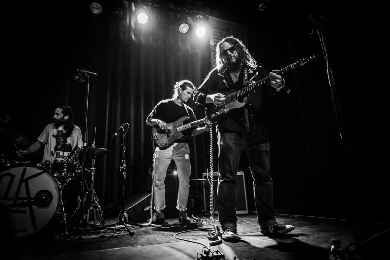 Een muzikant speelt gitaar op het podium, terwijl twee andere bandleden hun instrumenten bespelen in een sfeervolle zwart-wit setting.