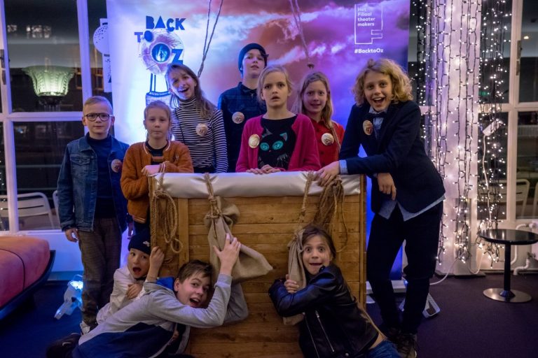 Een groep kinderen en volwassenen poseert rond een grote houten kist met een kleurrijke achtergrond.