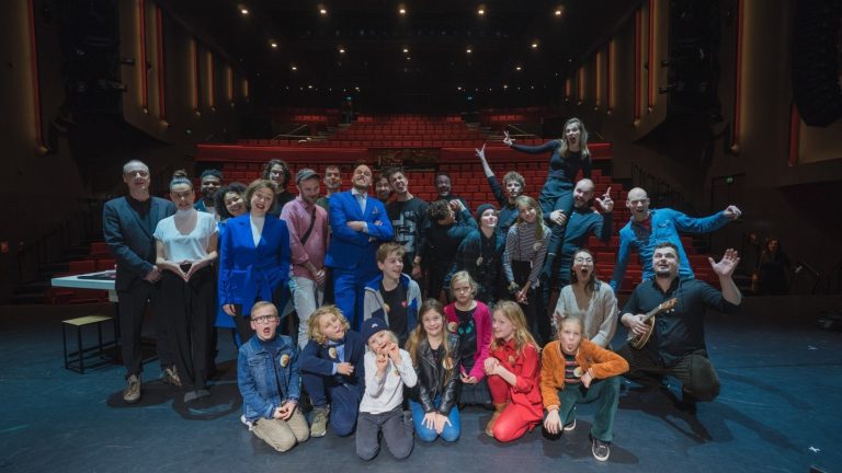 Een grote groep mensen poseert samen op het podium van een theater met rode stoelen op de achtergrond.