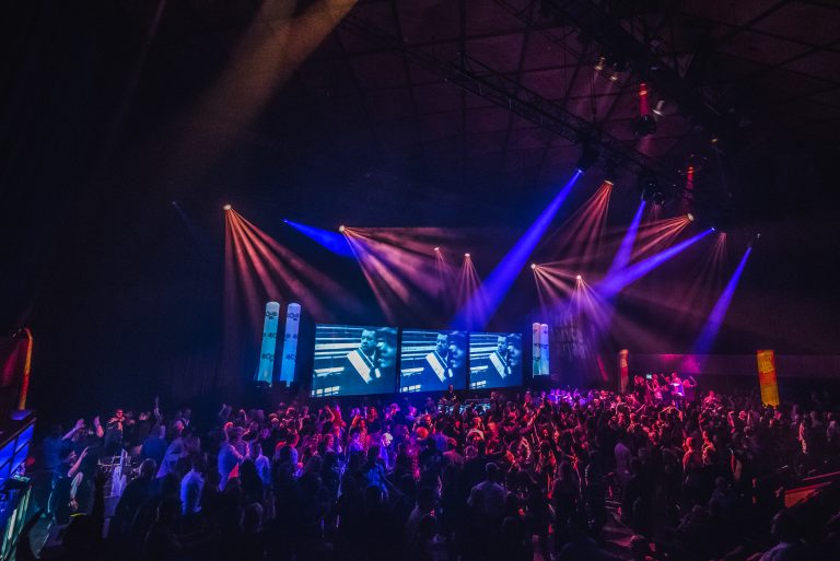 Een levendige muziekevenement met een menigte, gekleed in kleurrijke verlichting en videoprojecties op grote schermen.