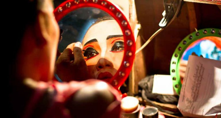 Een persoon die zich opmaakt in een spiegel met een rode rand, met opvallende make-up en kleurrijke ogen.