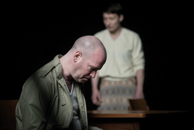 Twee mannen op een toneel, één met een kaal hoofd en een downcast houding, terwijl de ander in een lichte, casual outfit staat.