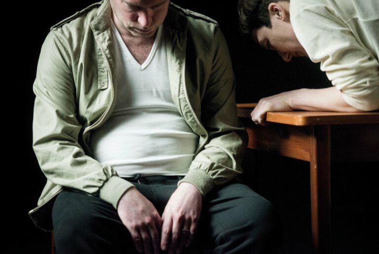 Twee mannen zitten in een sombere setting, één met zijn hoofd gebogen en de ander leunt op een tafel.