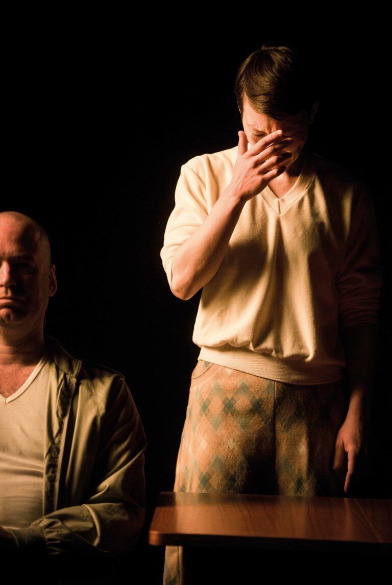 Twee mannen staan in dramatisch verlichte omgeving, waarbij één man zit en de ander emotioneel lijkt te zijn.