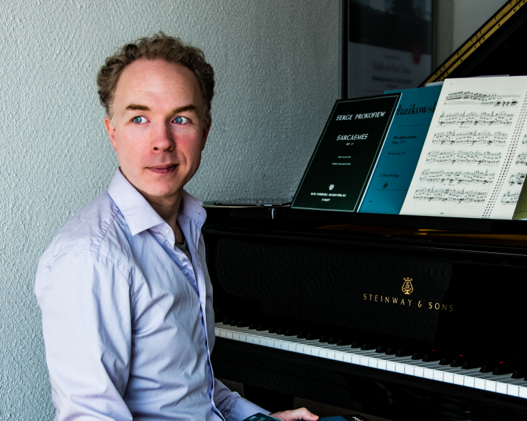 Een persoon zit naast een piano met bladmuziek in de tweede helft van het beeld.