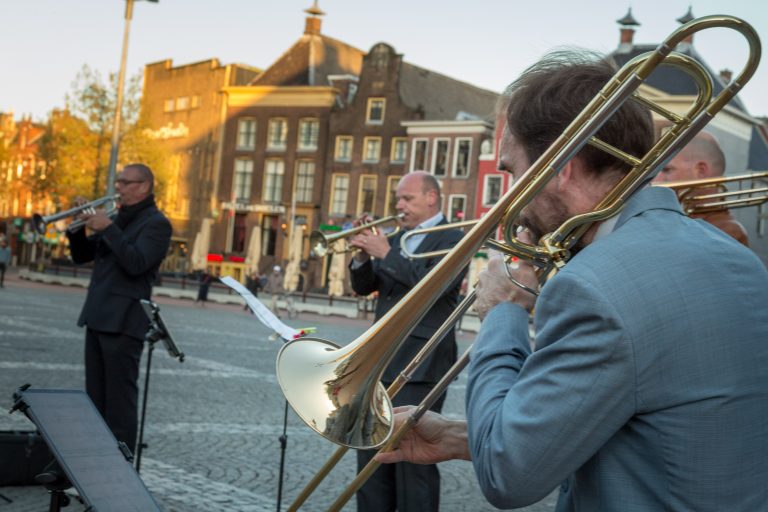 Drie muzikanten spelen op straat, met een focus op een trombonist, tegen een achtergrond van historische gebouwen in de avondzon.