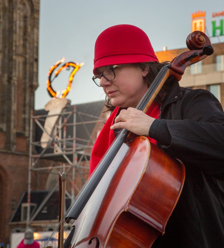 Een muzikant in een rode hoed speelt een cello op straat, met gebouwen op de achtergrond.