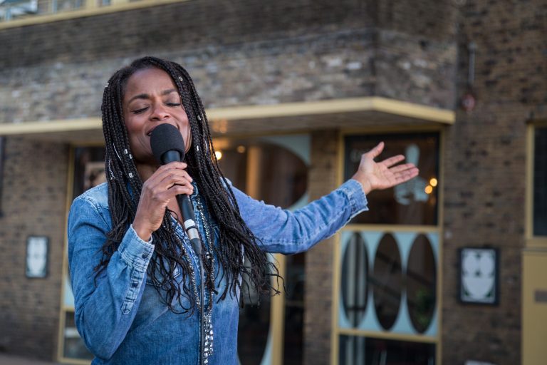 Een vrouw met lange vlechten zingt met een microfoon in handen, terwijl ze haar andere hand uitnodigend uitsteekt.