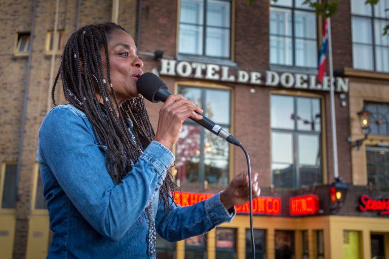 Zangeres met een microfoon zingt voor een hotel, omringd door een levendige sfeer.