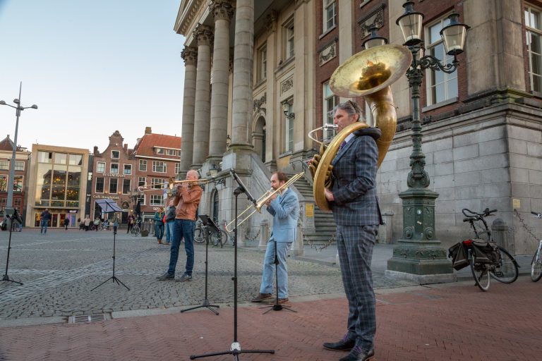 Een muzikale groep met een tuba, trombone en een andere blaasinstrument speelt buiten bij een historisch gebouw.