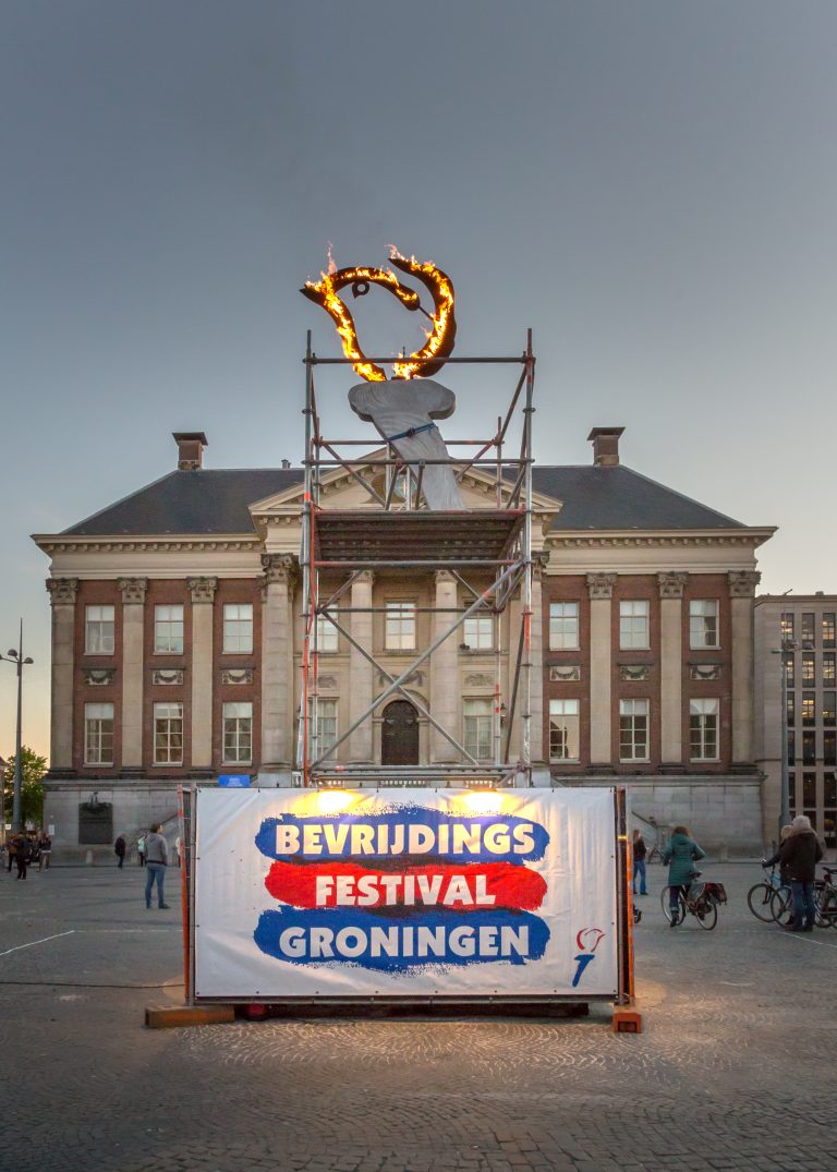 Een grote banner voor het Bevrijdingsfestival Groningen, met op de achtergrond een statig gebouw en een kleurrijk kunstwerk op een steiger.